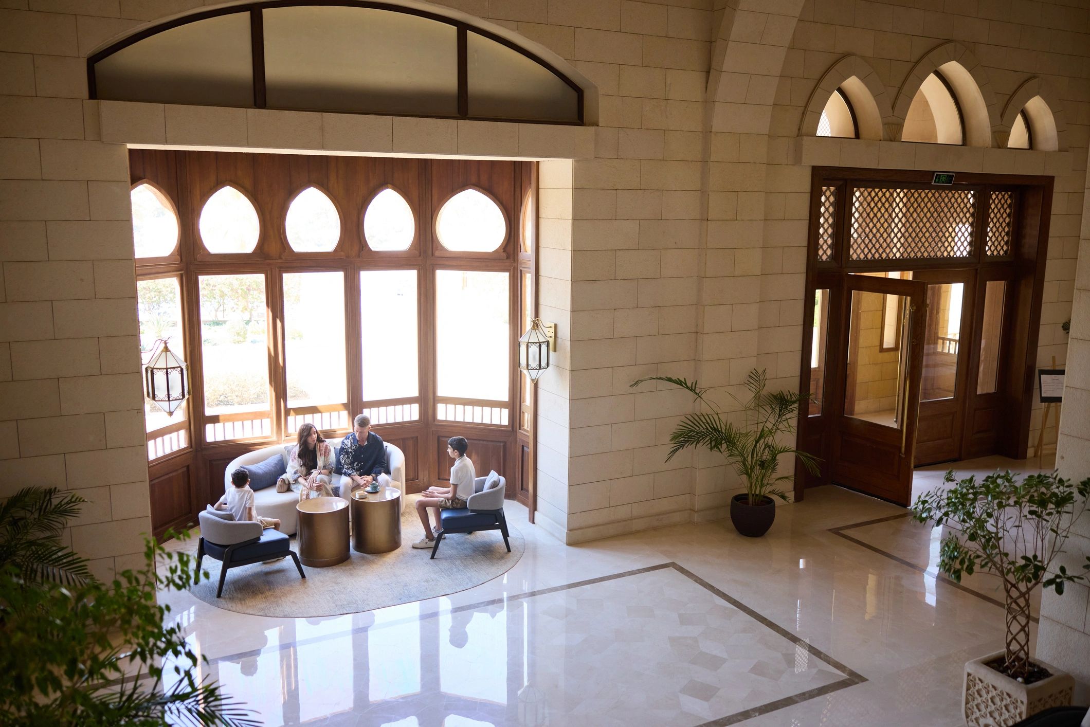 Family chatting in a sunlit hotel lobby seating area with arched windows and marble floor